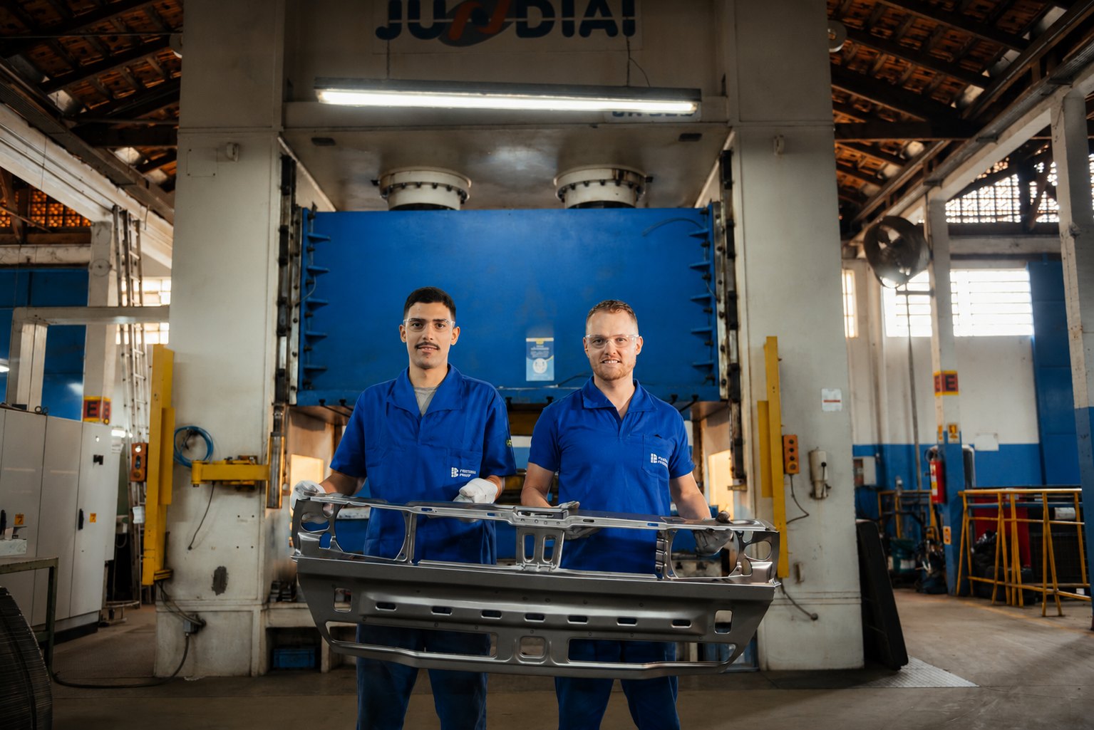 Dois colaboradores da Ponteiras Rodrigues, de uniforme azul e óculos de proteção, seguram um painel dianteiro estampado em aço à frente da prensa Jundiaí em Joinville/SC